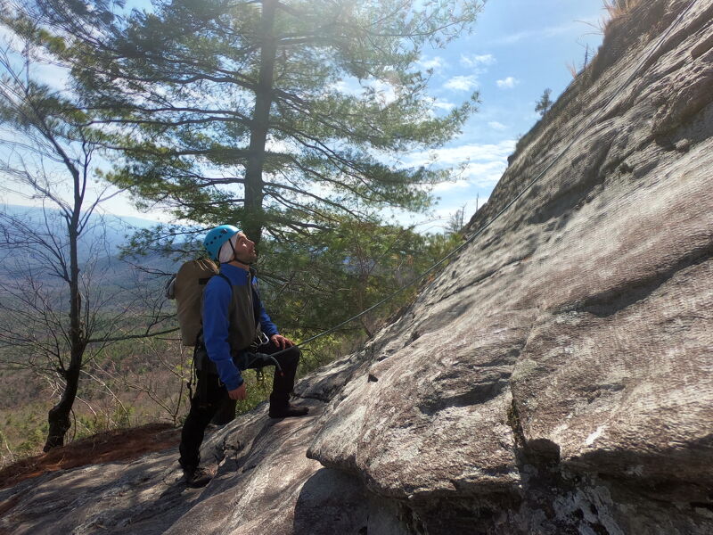 A person with a backpack and climbing gear stands on a rocky outcrop, looking up at a steep rock face. The individual is dressed in dark pants, a blue jacket, and a helmet. A tall pine tree grows nearby, and a forest stretches out in the background under a partly cloudy sky. The lighting suggests it's a sunny day, highlighting the textures of the rock and the surrounding landscape.